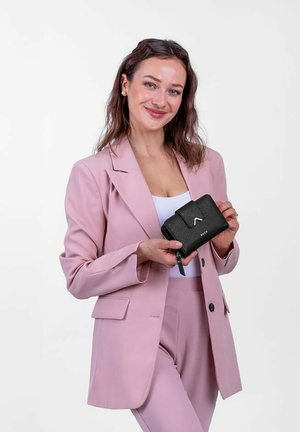 Black textured wallet with a zippered closure, featuring a small clasp and embossed logo, held by a person in a light pink blazer and pants.