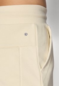 Cream-colored sweatpants with a textured surface, featuring side pockets and a small gray logo accent on the waistband.