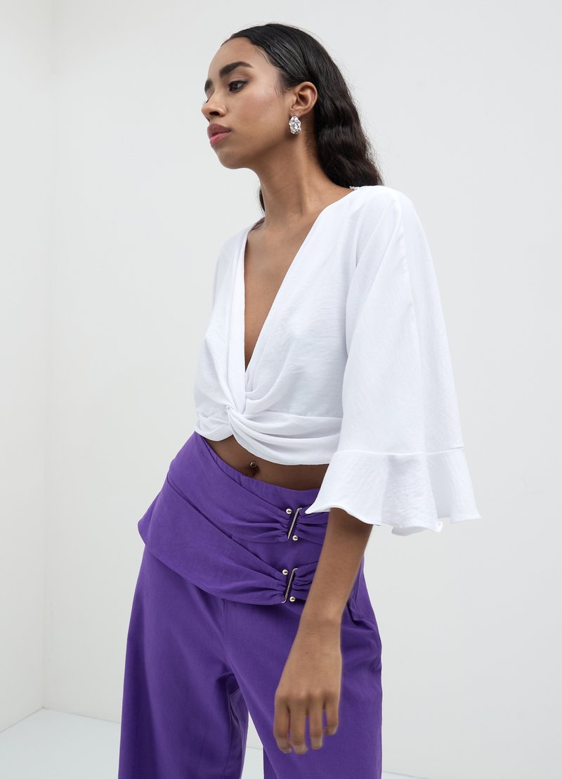 Crop top blanc avec un nœud à l'avant et des manches évasées, associé à un pantalon violet taille haute avec un design drapé et des accents métalliques.