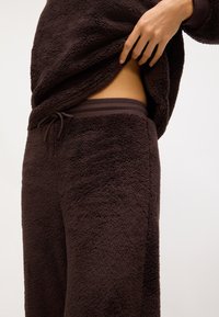 Das braune Fleece-Loungewear-Set besteht aus einem weichen, strukturierten Stoff. Das Oberteil hat eine lockere Passform und einen gerippten Bund am unteren Ende. Die Hose hat einen verstellbaren Bund.