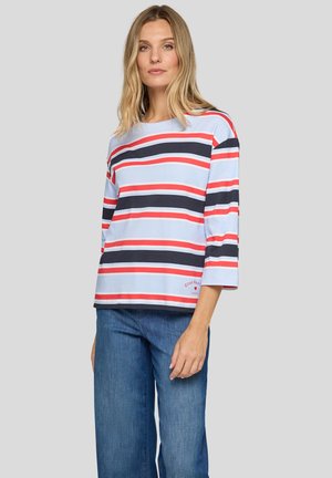Woman wearing a light blue, red, and navy striped long-sleeve shirt with "Good Vibes" text near hem, paired with blue jeans.
