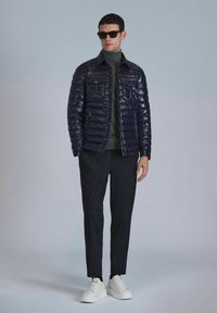 Veste matelassée navy avec finition brillante, quatre poches avant, col montant, assortie à un pantalon sombre et des baskets blanches.