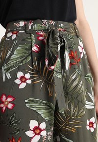 Green floral trousers featuring a tie waist, with a vibrant pattern of large leaves and flowers in red, pink, and white. Lightweight fabric.