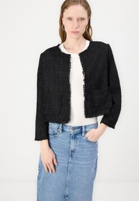 Svart cropped jakke i strukturert stoff med frynsede kanter, har en rund hals og to frontlommer. Kombinert med en hvit topp og denimskjørt.