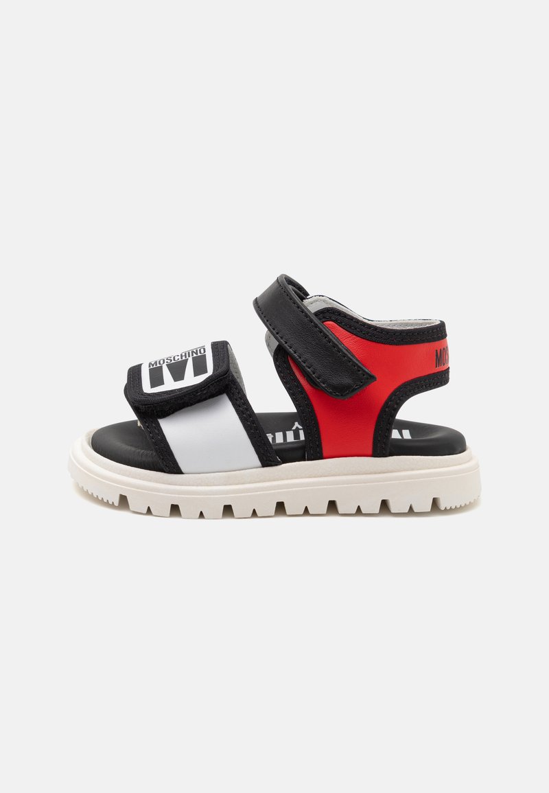 Sandalias negras, blancas y rojas con una correa de velcro, una suela blanca gruesa y un logo prominente en la correa superior; diseño deportivo.