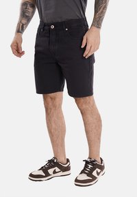 Shorts in denim nero con un taglio dritto, caratterizzati da una chiusura con bottone, passanti per la cintura e dettagli di cucitura sottili. Indossati con sneaker marroni e bianche.