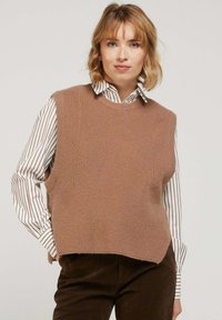 Pull sans manches en tricot beige avec un col rond, pr�ésentant une finition texturée et des fentes latérales, porté par-dessus une chemise rayée blanche et marron.