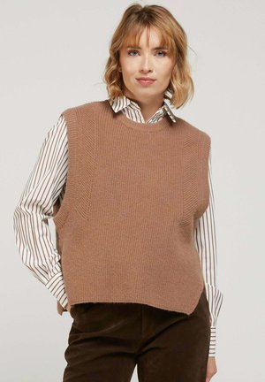 Pull sans manches en tricot beige avec un col rond, présentant une finition texturée et des fentes latérales, porté par-dessus une chemise rayée blanche et marron.
