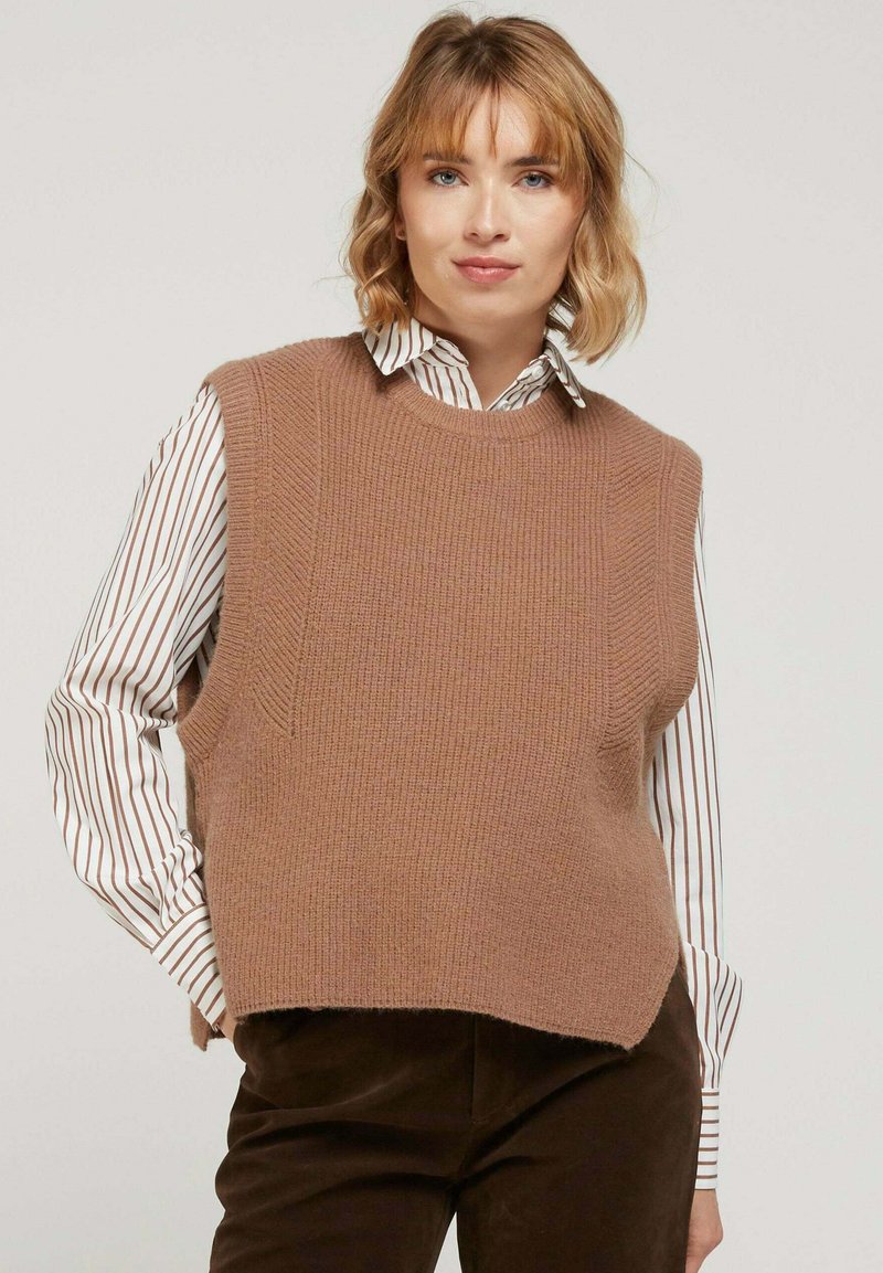 Pull sans manches en tricot beige avec un col rond, présentant une finition texturée et des fentes latérales, porté par-dessus une chemise rayée blanche et marron.