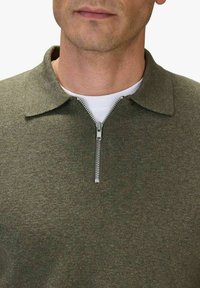 Olivgrön stickad polotröja med dragkedja vid kragen, strukturerad tyg och en vit t-shirt under, med fokus på en avslappnad design.