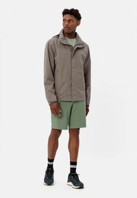 Grey waterproof jacket with a zip front, black accents, standing above knee-length green shorts. Black and white striped socks, dark shoes.