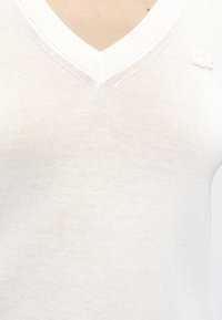Gros plan d’un pull blanc en maille à col en V, montrant la texture du tricot et un petit logo en relief sur la partie haute de la poitrine, près de l’épaule.