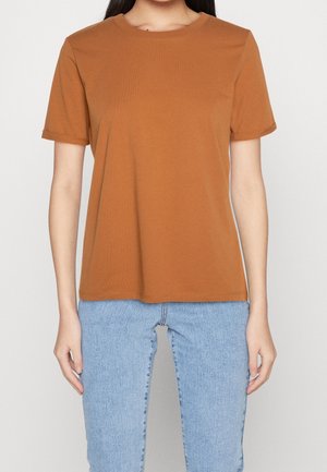 Kobieta w prostym brązowym t-shircie z krótkim rękawem i jasnoniebieskich dżinsach, stojąca na białym tle.