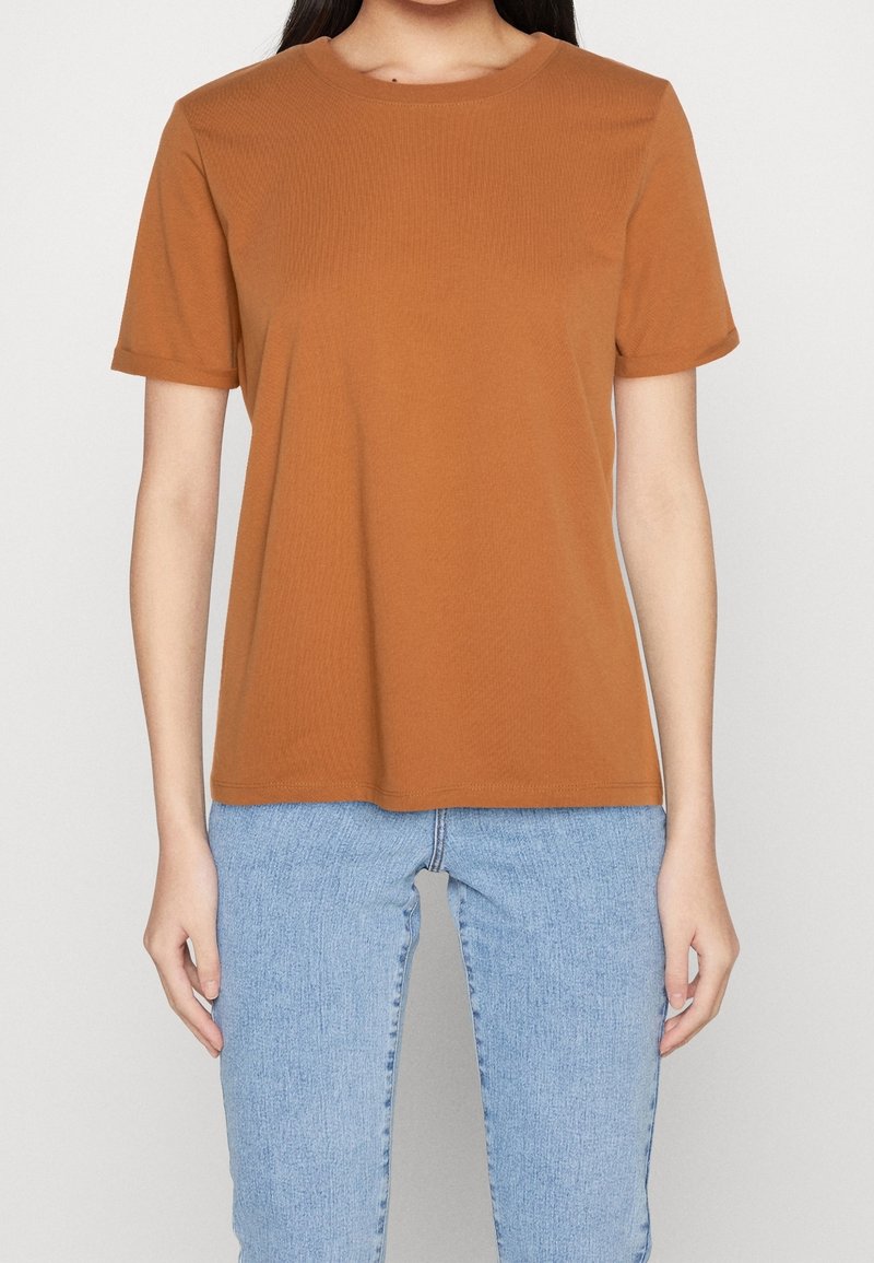 Femme portant un t-shirt marron uni à manches courtes et un jean bleu clair, debout devant un fond blanc.