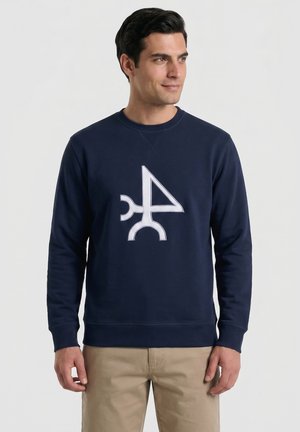 Hombre con sudadera azul marino con diseño geométrico abstracto blanco, de pie contra un fondo claro y sencillo.