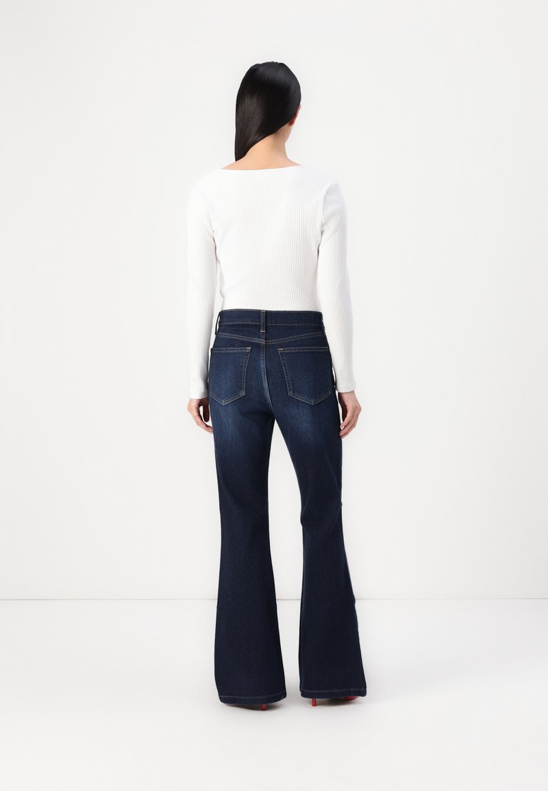 Jean bleu foncé évasé en denim, avec une taille haute, deux poches arrière et une finition nette aux ourlets. Associé à un top blanc à côtes.