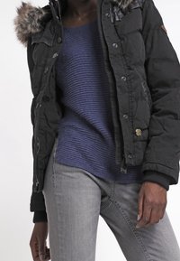 Veste noire zippée avec bordure en fausse fourrure, pull marine côtelé et jean gris. Tissu texturé, coupe ajustée, et accents en métal doré.