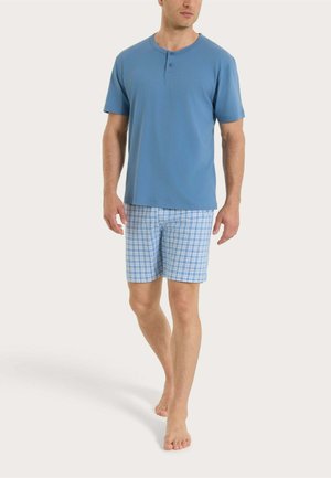 Uomo che cammina a piedi nudi, indossando una maglia henley a maniche corte blu e pantaloncini a quadretti blu e bianchi, su uno sfondo semplice.