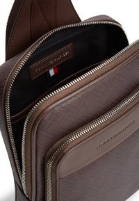 Καφέ δερμάτινη τσάντα με υφή στην εξωτερική πλευρά, πολλαπλές θήκες με φερμουάρ και λογότυπο. Εσωτερικά διαθέτει υφασμάτινη επένδυση.