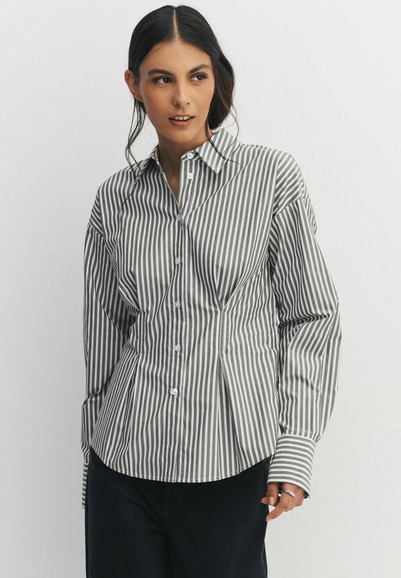 Woman wearing a fitted, long-sleeve, button-up shirt with vertical gray and white stripes, paired with dark pants, standing against a plain background.
