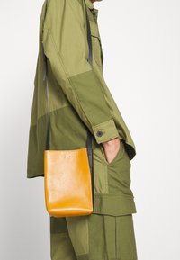 Borsa a tracolla gialla in pelle con una texture liscia, caratterizzata da una semplice forma rettangolare e una tracolla nera, esposta su un outfit verde oliva.