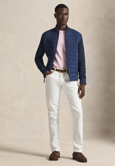 Μπλε καπιτονέ μπουφάν με ριμπ μανίκια, ανοιχτό ροζ μπλουζάκι, λευκό παντελόνι και καφέ παπούτσια σουέντ. Το μοντέλο στέκεται με τα χέρια στις τσέπες.