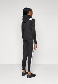 Zwarte sportieve hoodie met een capuchon en witte accenten, gecombineerd met zwarte broek met witte zijstrepen. De stof lijkt glad en goed aansluitend.