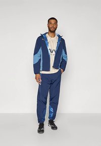 Mørkeblå vindjakke med lyseblå detaljer, glidelåslukking og hette, kombinert med matchende bukser. Brukt over en stripete lys skjorte.