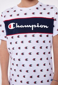 Cotton t-shirt featuring a white base with repeated small logo patterns in red and black, a navy horizontal band with "Champion" in white script.
