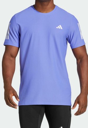 Mężczyzna w jasnofiolecie sportowej koszulce Adidas z krótkim rękawem i białym logo z trzema paskami na rękawach i piersi.