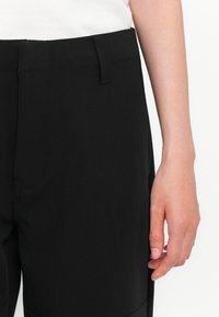 Zwarte broek met een zachte textuur, voorzien van een strakke tailleband en subtiele zijzakken, tentoongesteld met een hand ernaast.