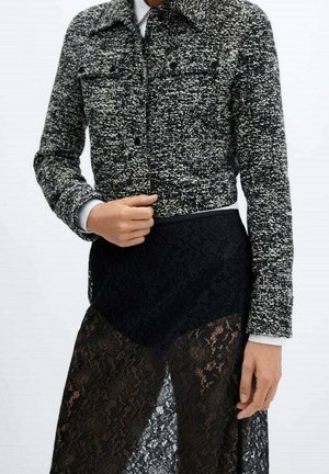 Femme portant une veste courte texturée noire et blanche sur une jupe en dentelle noire avec une superposition transparente, montrant la main ajustant la veste.