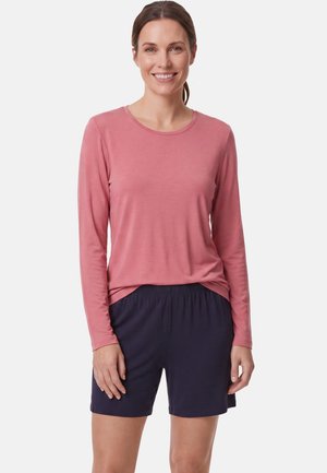 Lachende vrouw met bruin haar in een paardenstaart, draagt een roze top met lange mouwen en donkerblauwe korte broek, staand tegen een effen lichte achtergrond.