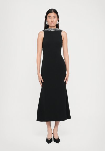 Schwarzes ärmelloses Kleid mit tailliertem Oberteil und ausgestellt Rock, das einen mit Perlen besetzten Ausschnitt und eine subtile Textur aufweist. Schwarze spitz zulaufende High Heels.
