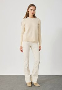 Beige stickad tröja med rund halsringning och ribbad fåll, kombinerad med vita vida byxor och beiga skor. Minimalistisk design och textur.