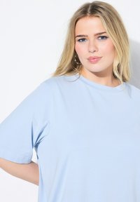 Světle modré bavlněné tričko s krátkým rukávem, kulatým výstřihem a hladkou texturou. Model pózuje na neutrálním pozadí.