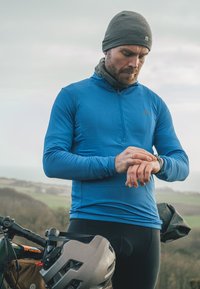 Blauwe sportshirt met lange mouwen en ritskraag, gedragen onder een grijze beanie. Man die op zijn horloge kijkt terwijl hij naast een fiets staat met een helm.