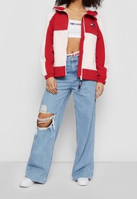 Czerwona i kremowa kurtka przeciwwiatrowa z kapturem, zapinana na zamek, z elastycznymi mankietami. Jasnoniebieskie szerokie, przetarte dżinsy. Białe sneakersy.