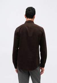 Brązowa koszula z długim rękawem o prostym wykończeniu i guziczkami przy mankietach, z plisą na plecach dla swobody ruchów, wykonana z teksturowanej tkaniny.