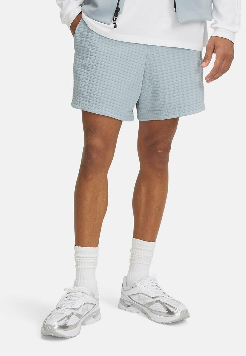 Pantalones cortos de un azul claro con textura que presentan un patrón acolchado, bolsillos laterales y un corte relajado, combinados con zapatillas deportivas blancas y calcetas.
