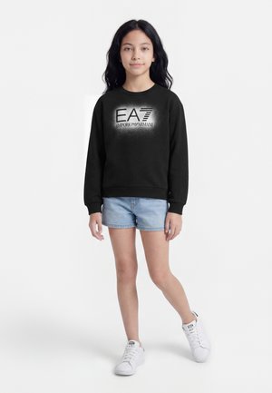 Djevojka s dugom crnom kosom, odjevena u crnu Emporio Armani majicu s kapuljačom, svijetle traper kratke hlače i bijele tenisice, stoji na bijeloj pozadini.