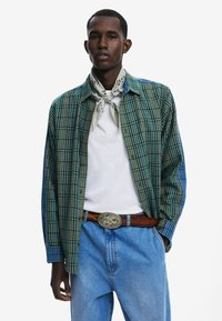 Camisa de cuadros en tonos verdes y azules, con diseño de botones. Combinada con una camiseta blanca y jeans de mezclilla, accessoriza con una bufanda estampada.