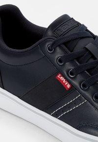 Sapatilha de couro azul marinho com sola branca, apresentando atacadores redondos, ilhós de metal e um logótipo vermelho da Levi's na lateral. Textura suave.