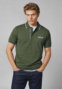 Mörkgrön polo skjorta med en vit randig krage, prydd med en knäppt placket och 'Red Bull Racing'-logga på bröstet. Bärs med mörka jeans.