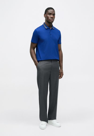 Mėlyna polo marškinėliai su dviem mygtukais ir geltonomis dryžuotomis apykaklėmis, derinami su pilkomis tekstūruotomis kelnėmis ir baltais sportbačiais, stovintis modelis.