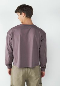 Langärmliges, kurzes Hemd aus einem gedämpften lila Stoff mit rundem Ausschnitt und lockerer Passform; kombiniert mit olivgrünen Cargo-Hosen.