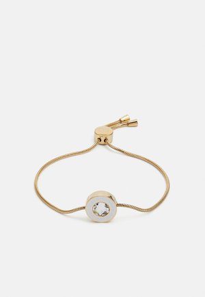 Bracciale regolabile in tonalità oro con un pendente circolare, dotato di un centro in cristallo trasparente e accenti bianchi attorno al logo. Design della catena testurizzata.