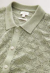 Ljusgrön stickad pikétröja med geometriskt spetsmönster, krage och två vita knappar. Etiketter synliga inuti kragen.