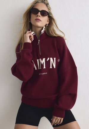 Blonde Frau mit schwarzen Sonnenbrillen, einem burgunderfarbenen Pullover mit halbem Reißverschluss und weißer Beschriftung sowie schwarzen Shorts, die vor einer schlichten weißen Wand posiert.