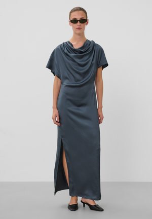 Femme portant une robe maxi drapée bleu foncé avec une fente côté, des escarpins noirs et des lunettes de soleil noires étroites sur un fond gris uni.